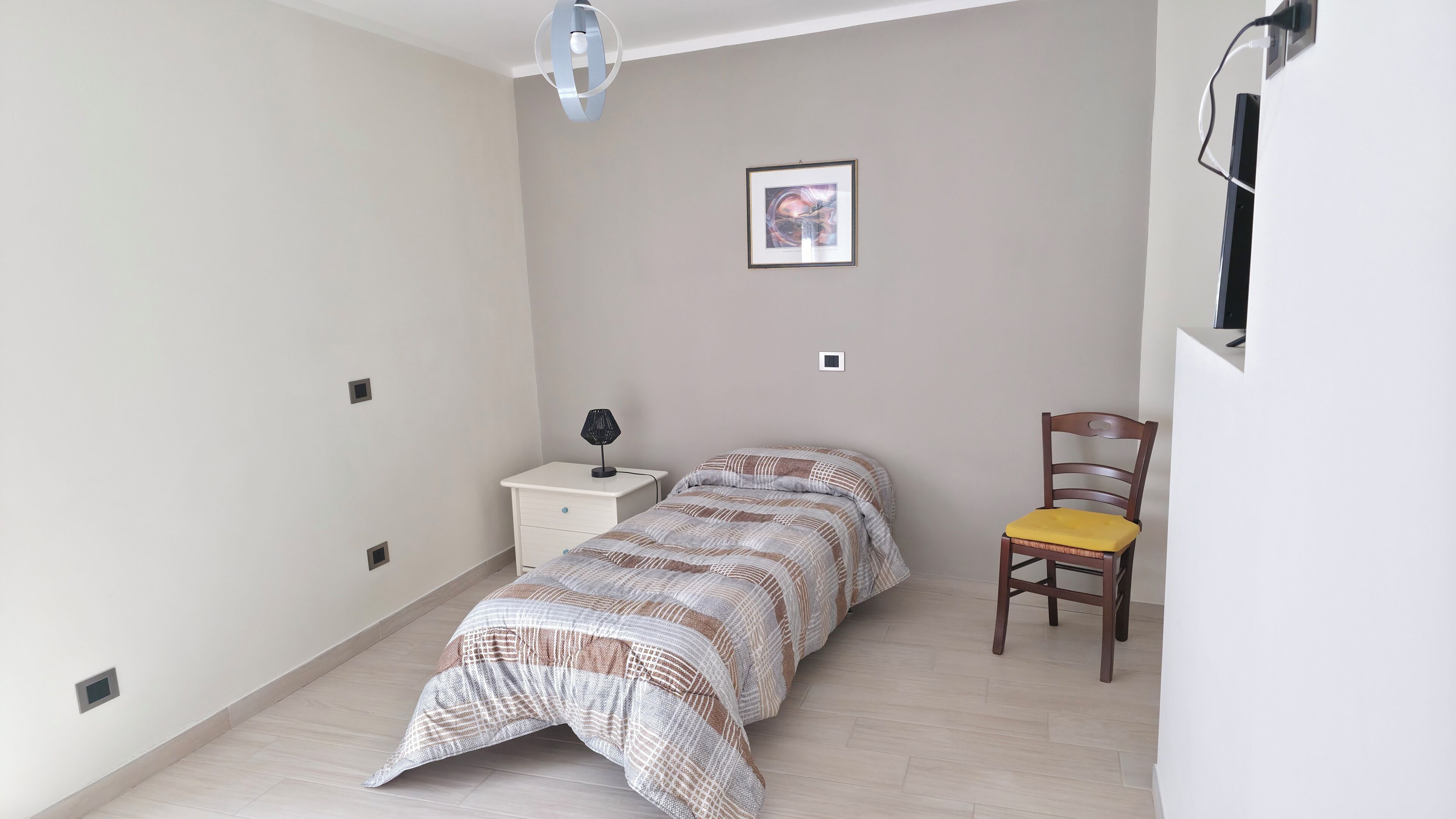A bedroom with a bed, a bedside table with a lamp, a chair, and a wall-mounted TV.