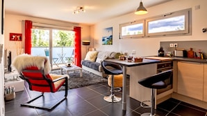 A modern kitchen with a bar area, large windows, and a cozy seating area with a red chair and a sofa. - Le Coquelicot (Annecy)