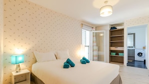 A neatly made bed with white linens and two rolled towels, a bedside lamp, and a wardrobe with open doors revealing shelves and a washing machine. - Le Coquelicot (Annecy)