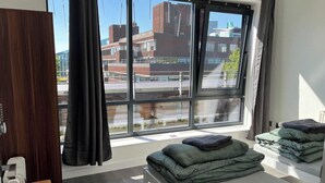 Ett hotellrum med ett stort fönster, en säng med grönt sänglinne, en garderob i trä och ett litet bord.