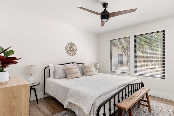 Un dormitorio con cama, ventilador de techo, mesita de noche con lámpara, planta en maceta y ventana con persianas.