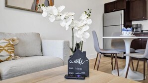 Ein modernes Wohnzimmer mit einer weißen Orchideen-Vase auf einem Holztisch, einem Kreidetafel-Schild mit WLAN-Informationen, einer Couch und einer Küche im Hintergrund.