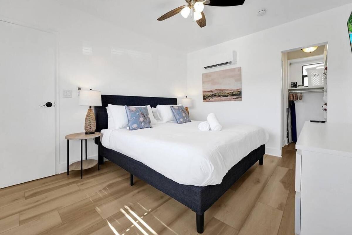 Un dormitorio con cama, mesita de noche, lámpara, ventilador de techo y una obra de arte en la pared.