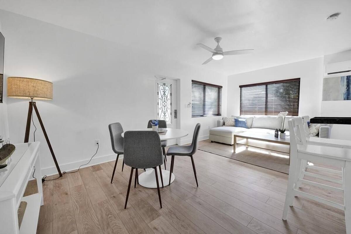 Una sala de estar moderna con comedor, paredes blancas, piso de madera, ventilador de techo y ventanas grandes con persianas.