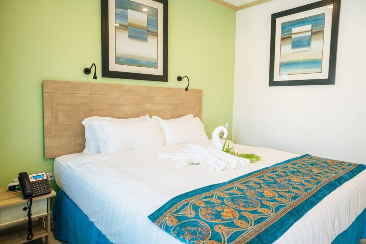 Una habitación de hotel con cabecera de madera, cama con sábanas blancas, colcha azul y dorada, teléfono sobre una mesita de noche y dos cuadros en la pared.