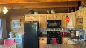 A cozy kitchen with wooden cabinets, a black refrigerator, a microwave, and a granite countertop. There are bar stools and a dining table with chairs.