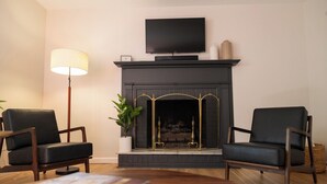 Una sala de estar con chimenea, dos sillones, una lámpara de pie y un televisor montado sobre la chimenea.