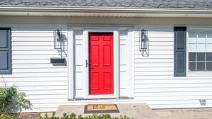 Una casa blanca con una puerta roja, dos lámparas de estilo farol y un felpudo con la inscripción 'OH HELLO'.