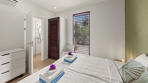 Ein modernes Schlafzimmer mit Bett, Nachttisch, Kommode, Dusche und einem Fenster mit Jalousien.