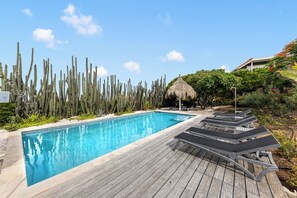 A swimming pool with wooden deck and lounge chairs, surrounded by cacti and tropical plants. - Ocean View Apartment Bayside, 6 guests - Boca Gentil, Jan Thiel area (Jan Thiel)