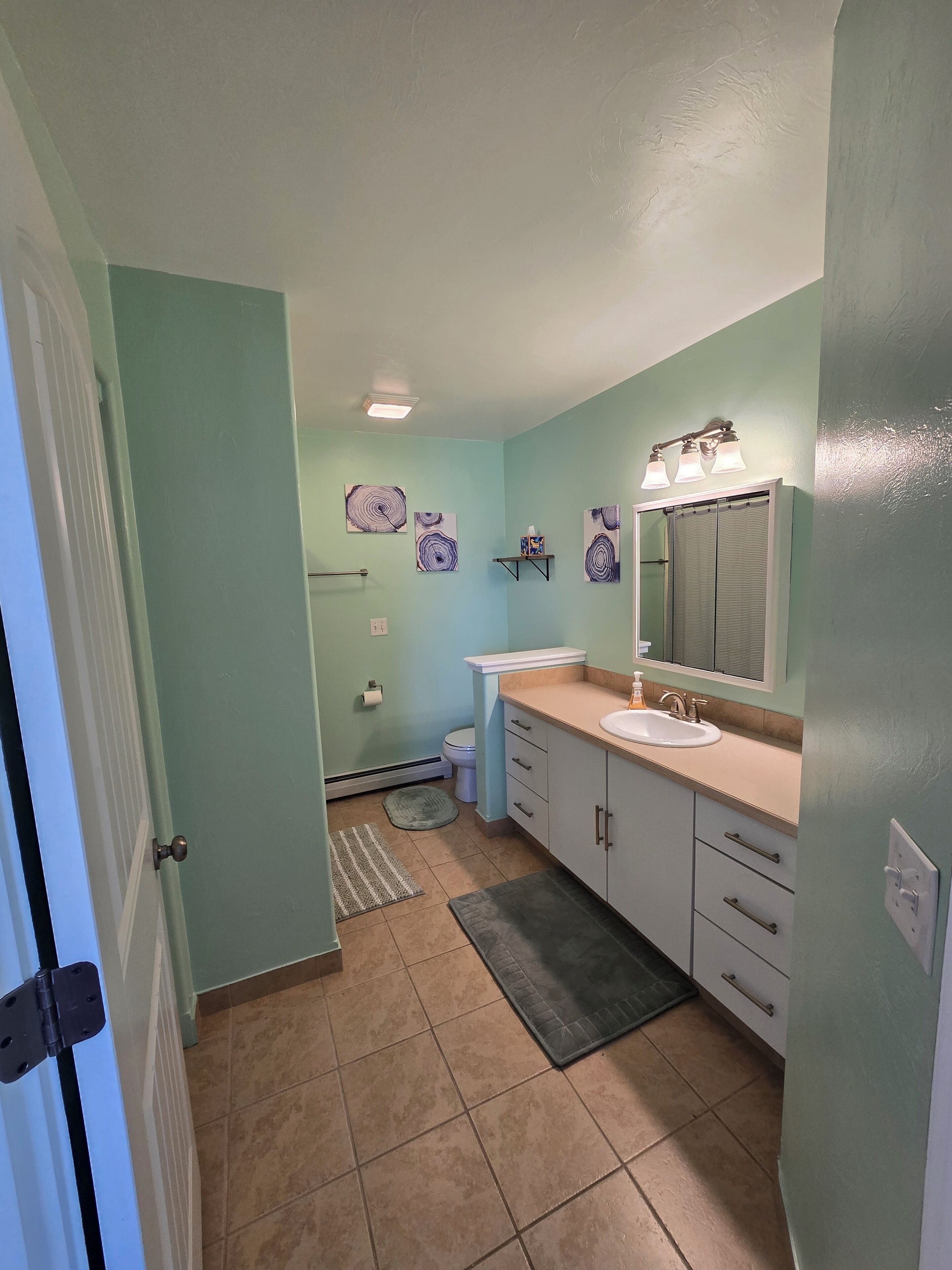 A bathroom with light blue walls, a white sink, a mirror, and a toilet. There are three wall-mounted lights and a small shelf with a box.
