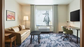 Habitación de hotel con sofá, una mesita, una silla, escritorio, televisor y ventana con cortinas.
