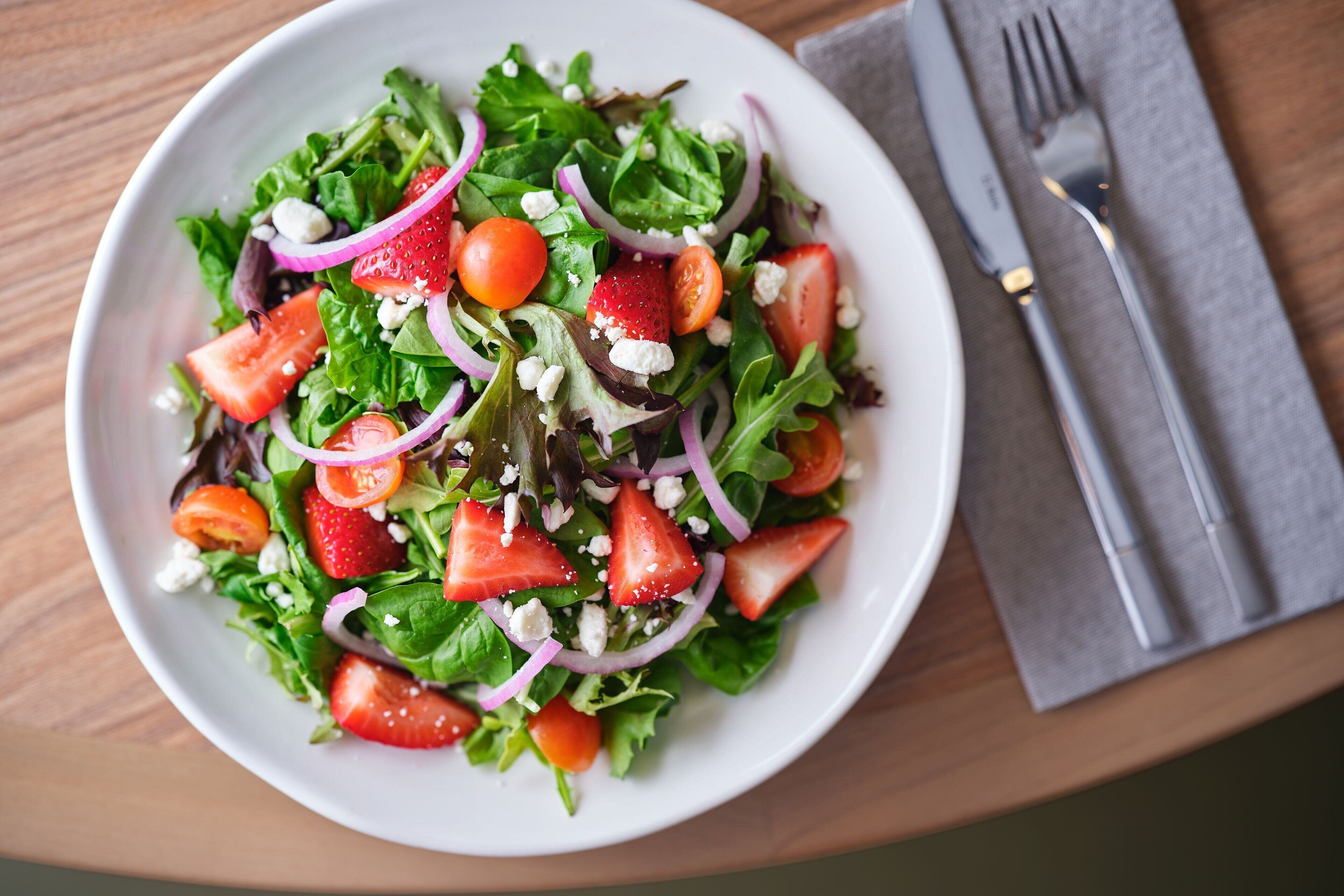 A white bowl of salad with mixed greens, strawberries, red onions, and feta cheese on a wooden table.