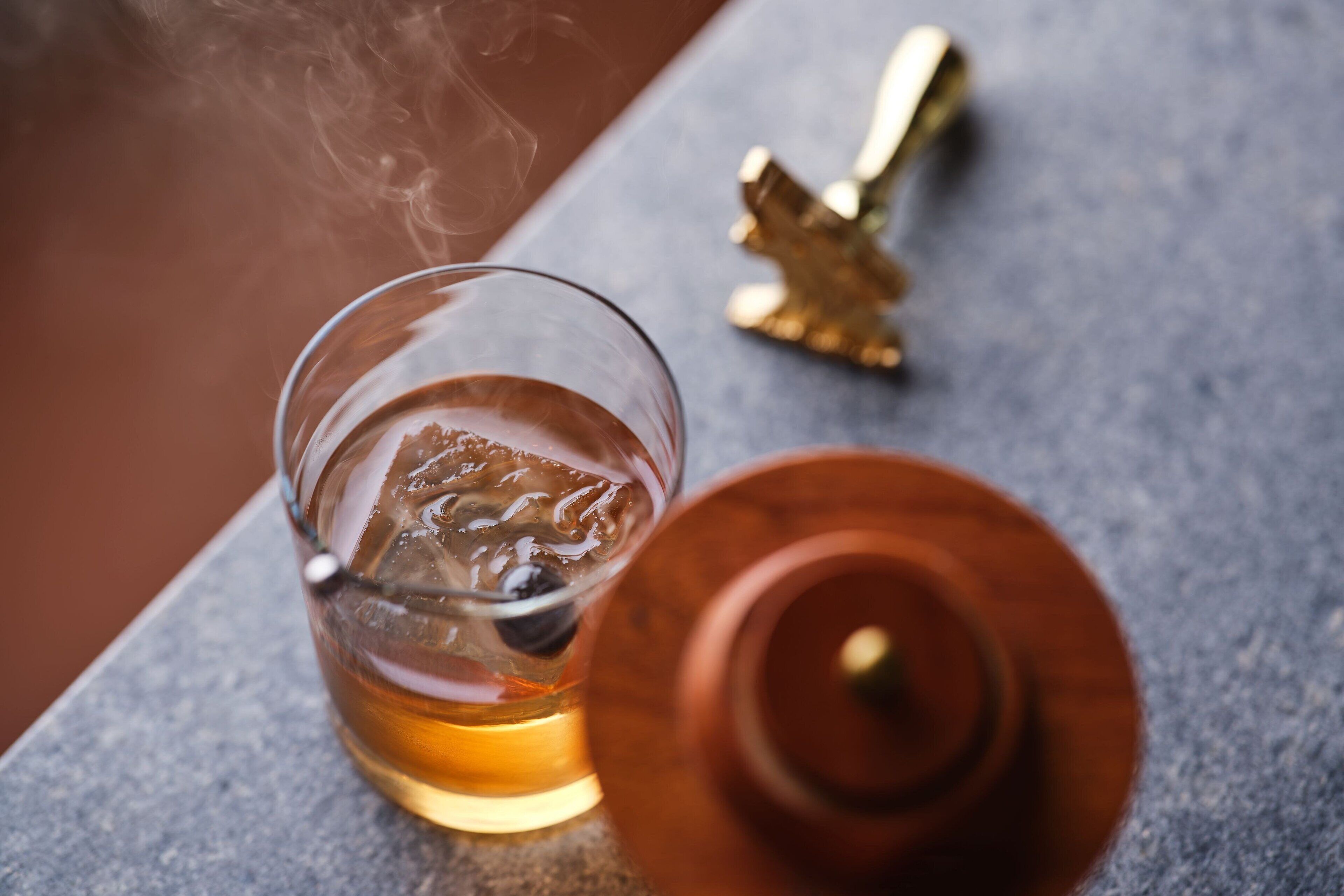 A glass of amber liquid with ice cubes, a golden object resembling a cross, and a brown container on a textured surface.