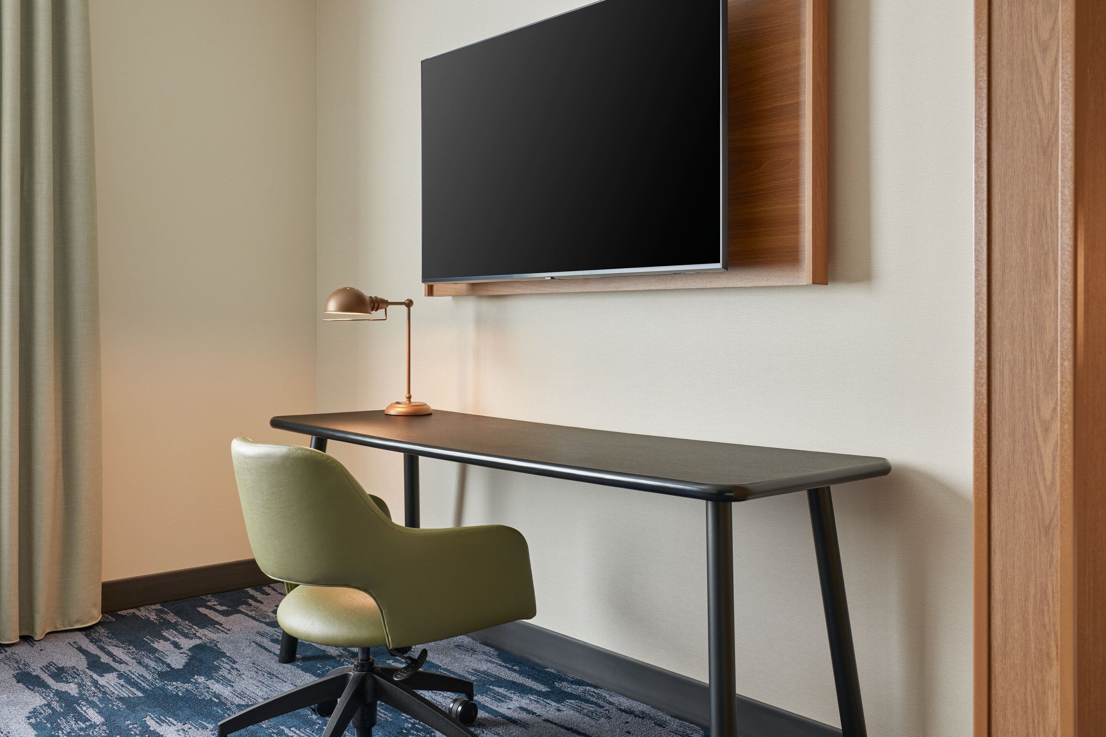 Quarto de hotel com escrivaninha, cadeira, televisão fixada na parede e abajur na escrivaninha.