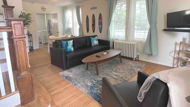 Un vaste salon avec un canapé noir, une table basse en bois et un téléviseur. On aperçoit un coin repas au fond et un escalier menant à un autre niveau.