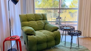 Un fauteuil vert, un tabouret rouge, un lampadaire noir et une table à motifs zébrés dans une pièce dotée de grandes fenêtres.