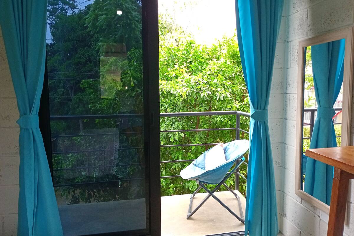 A balcony with a blue chair, a wooden table, and a view of greenery and trees.