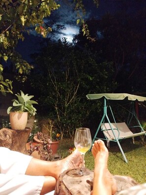 A person relaxing outdoors at night with a glass of drink, surrounded by greenery and a swing chair. - M&K House (La Banda de Shilcayo)
