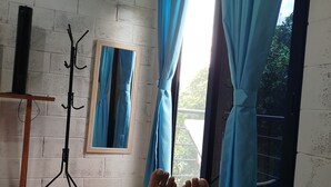 A room with a bed, a window with blue curtains, a coat rack, and a framed mirror.