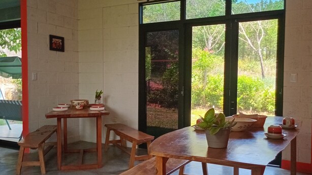 A dining area with wooden tables and benches, large windows with a view of greenery, and a hanging light fixture.