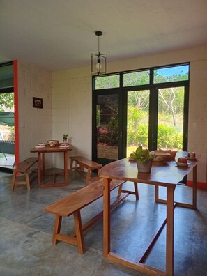 A dining area with wooden tables and benches, large windows with a view of greenery, and a hanging light fixture. - M&K House (La Banda de Shilcayo)