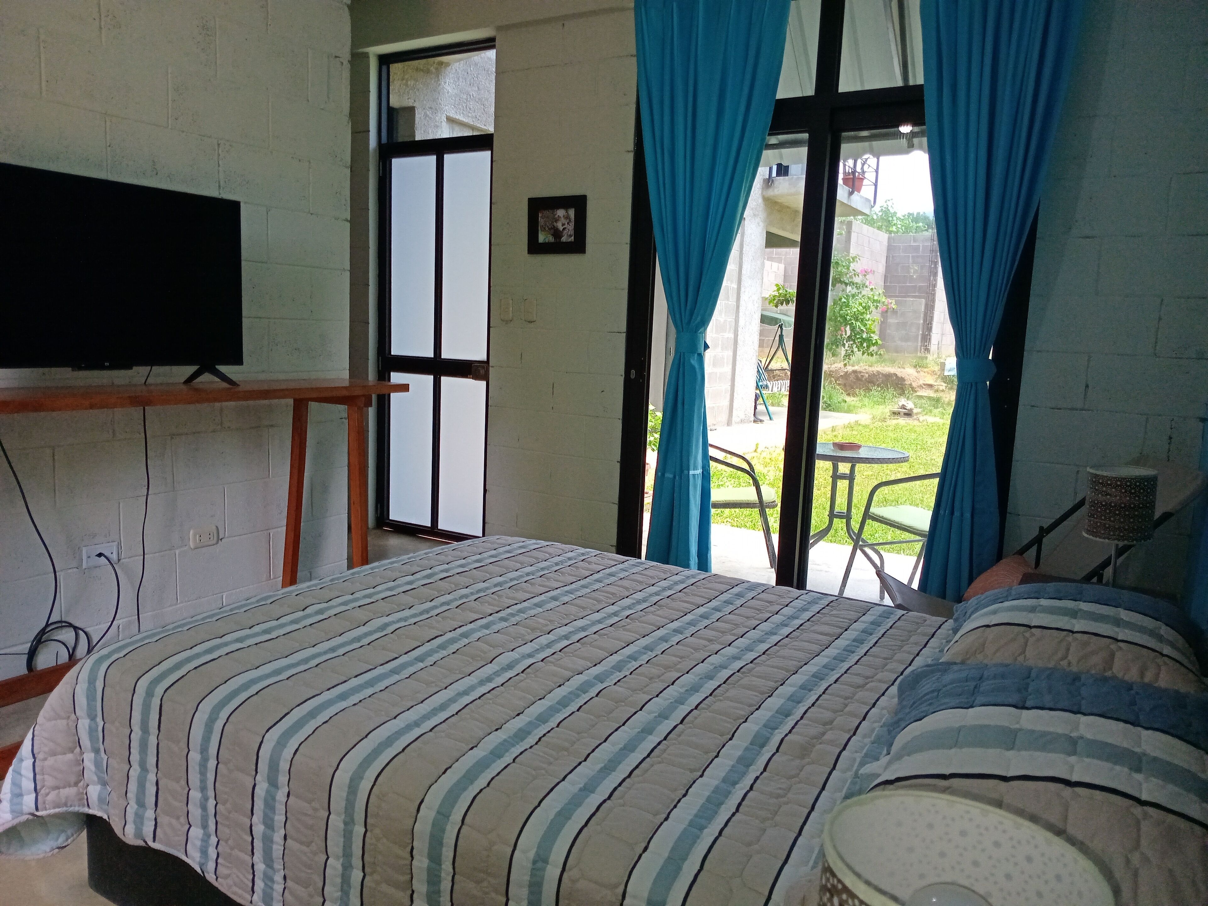 Un dormitorio con cama, televisor en un soporte, ventana con cortinas y vista al exterior.