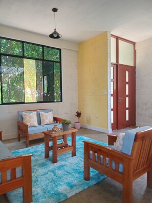 A room with a sofa, two wooden chairs, a coffee table, a blue rug, a potted plant, and a large window with a view of trees. - M&K House (La Banda de Shilcayo)