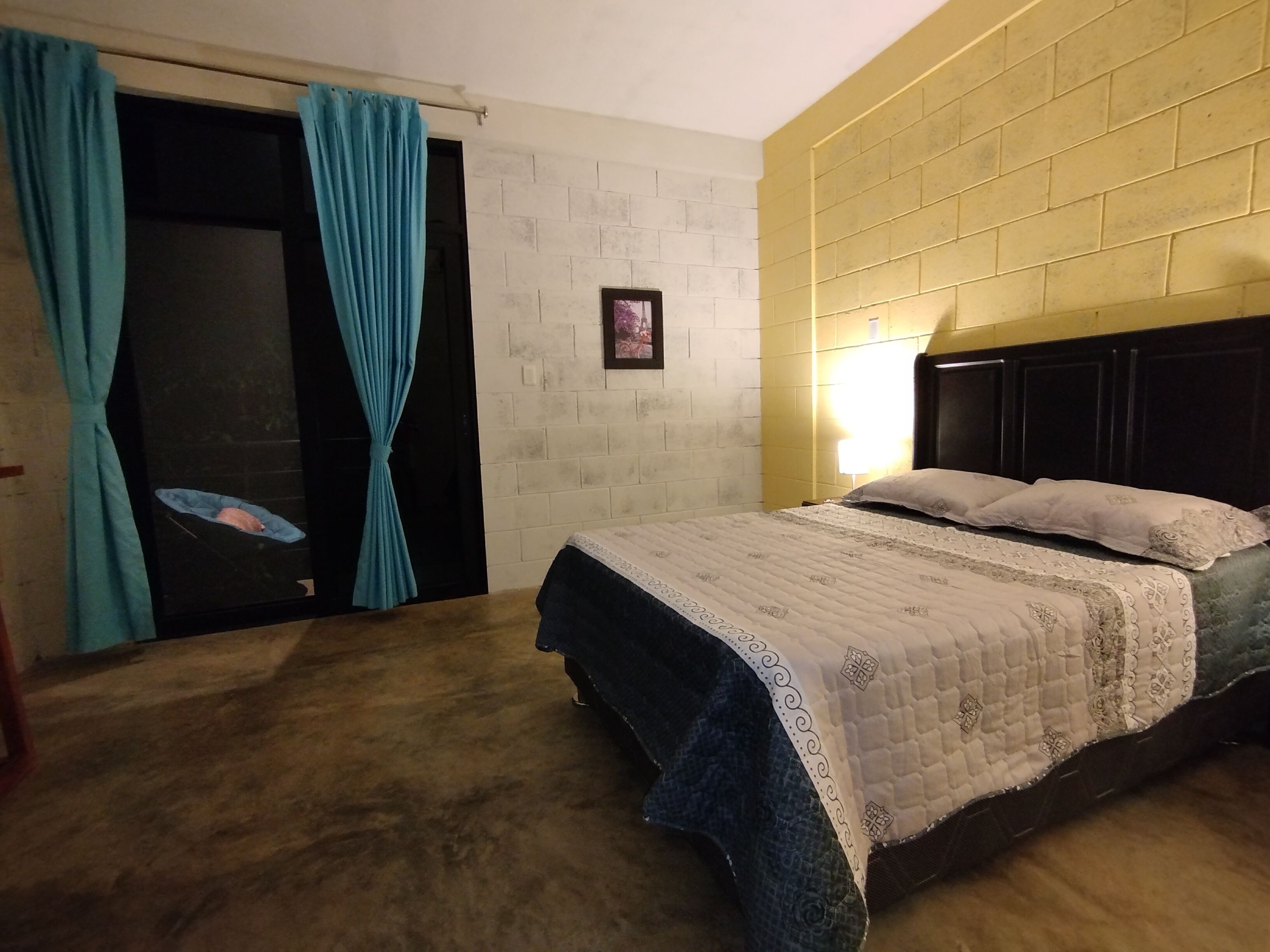 A bedroom with a bed, curtains, a wall-mounted light, and a framed picture.