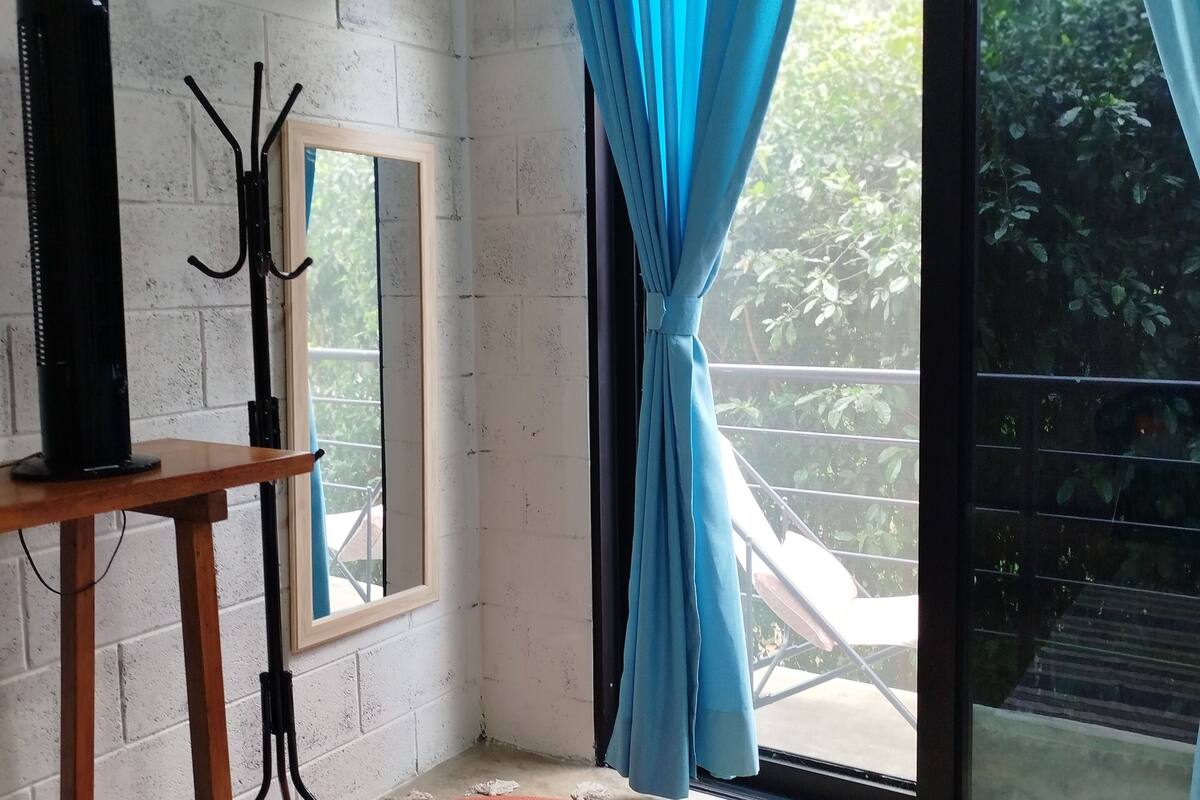 A room with a white brick wall, a wooden table with a black object on it, a mirror, a blue curtain, and a red rug.