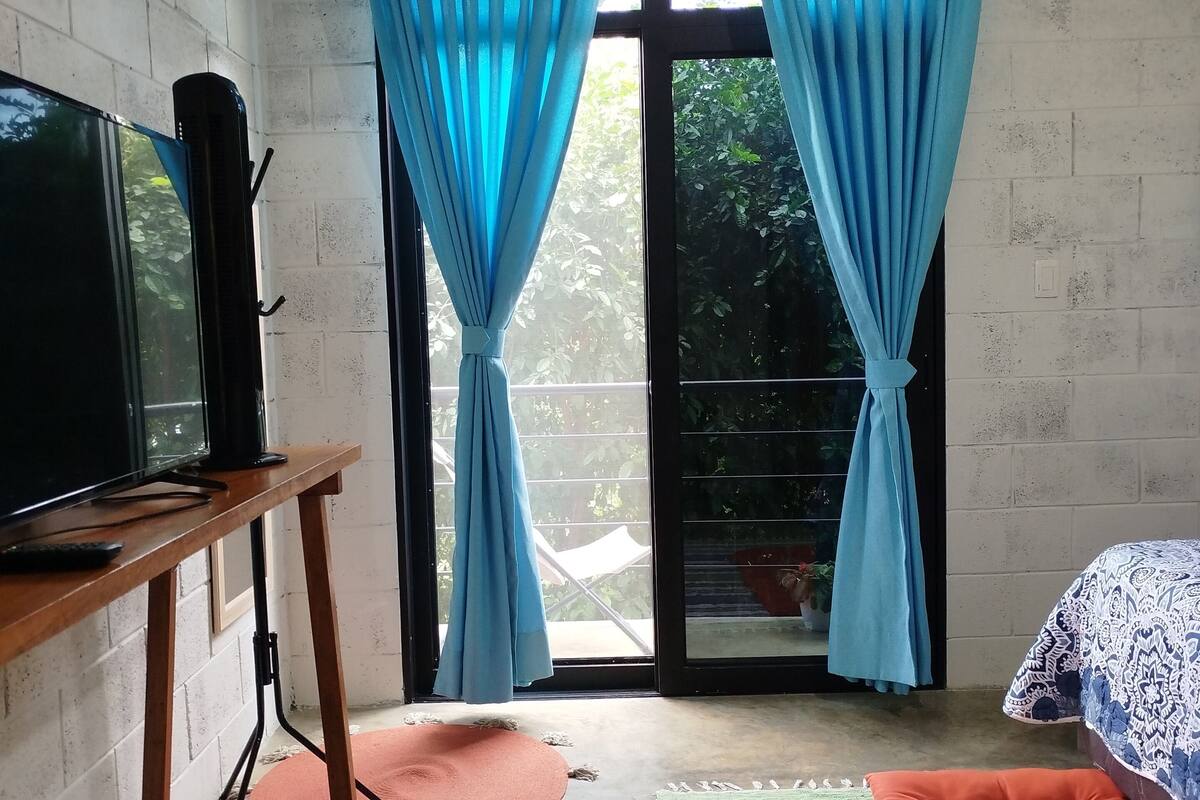 A room with a TV on a wooden stand, a bed with patterned bedding, a colorful striped rug, and blue curtains over a sliding glass door leading to an outdoor area.