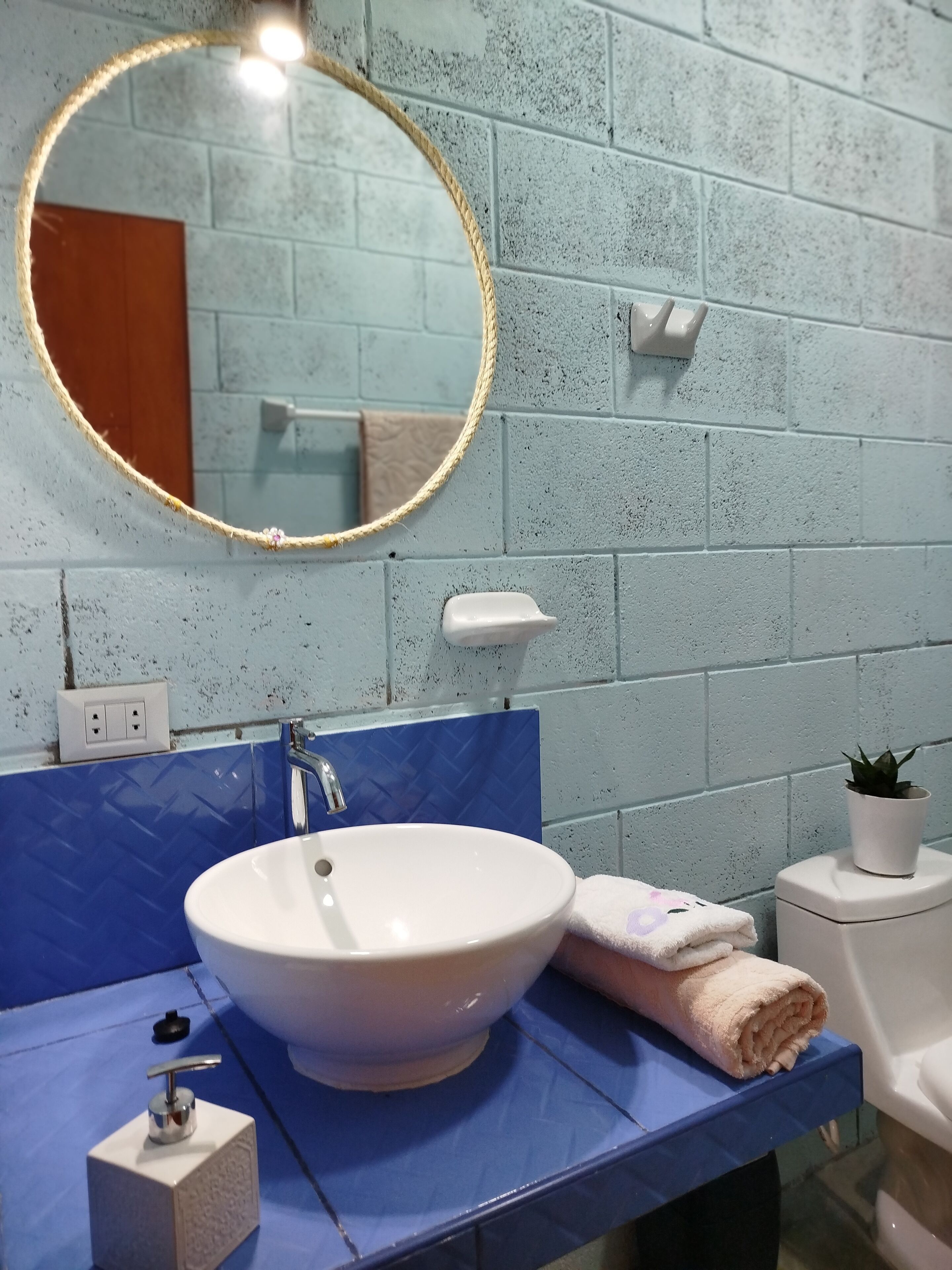 Un baño con lavamanos blanco, encimera azul y espejo redondo con marco de cuerda.