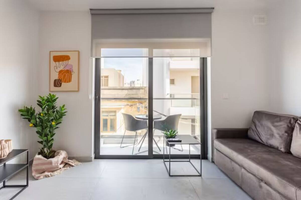 A modern living room with a sofa, a dining table with chairs, a potted plant, and a sliding glass door leading to a balcony.