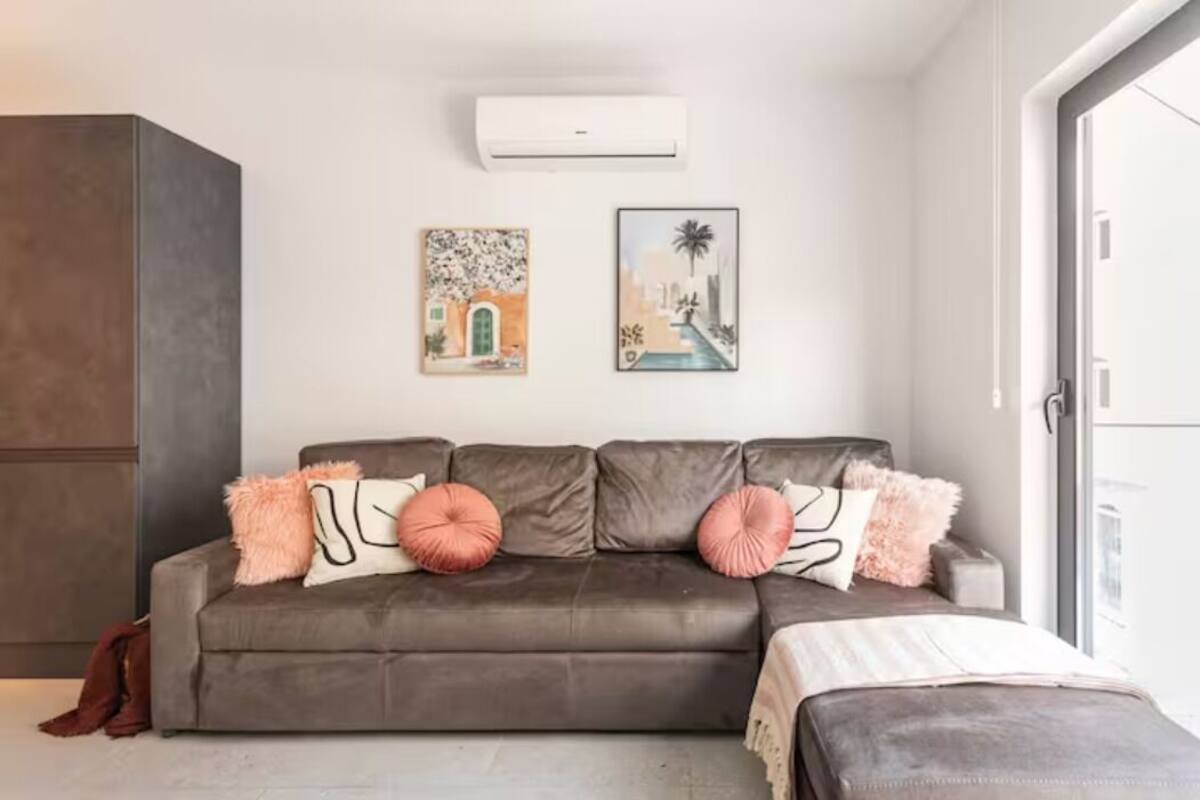 A modern living room with a grey sofa, two wall-mounted air conditioners, and two framed artworks depicting architectural scenes.