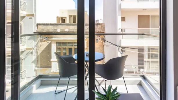 Ein Balkon mit Glasgeländer, zwei schwarze Stühle, ein kleiner Tisch und ein Blumentopf.