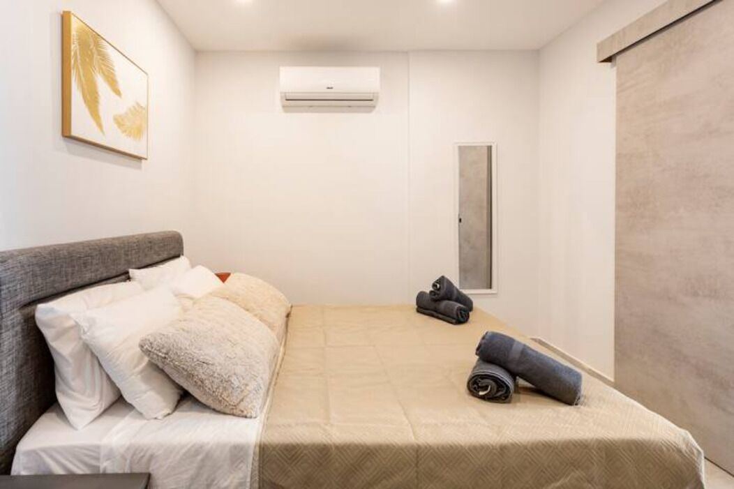 A modern bedroom with a bed, pillows, a wall-mounted air conditioner, and a framed artwork.