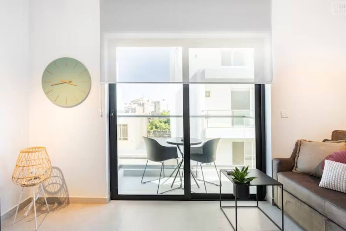 A modern living room with a large sliding glass door leading to a balcony, a round wall clock, a small table with a plant, and a sofa with cushions.