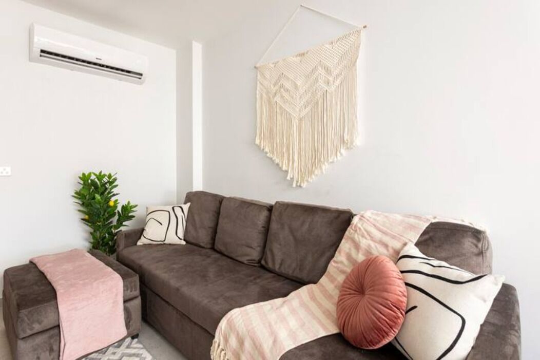Een moderne woonkamer met een grijze bank, een macramé wanddecoratie en een kleine potplant.