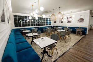 A restaurant with a long blue bench seating area, tables, and chairs. - Vasaka Maison Bandung (Sumur Bandung)
