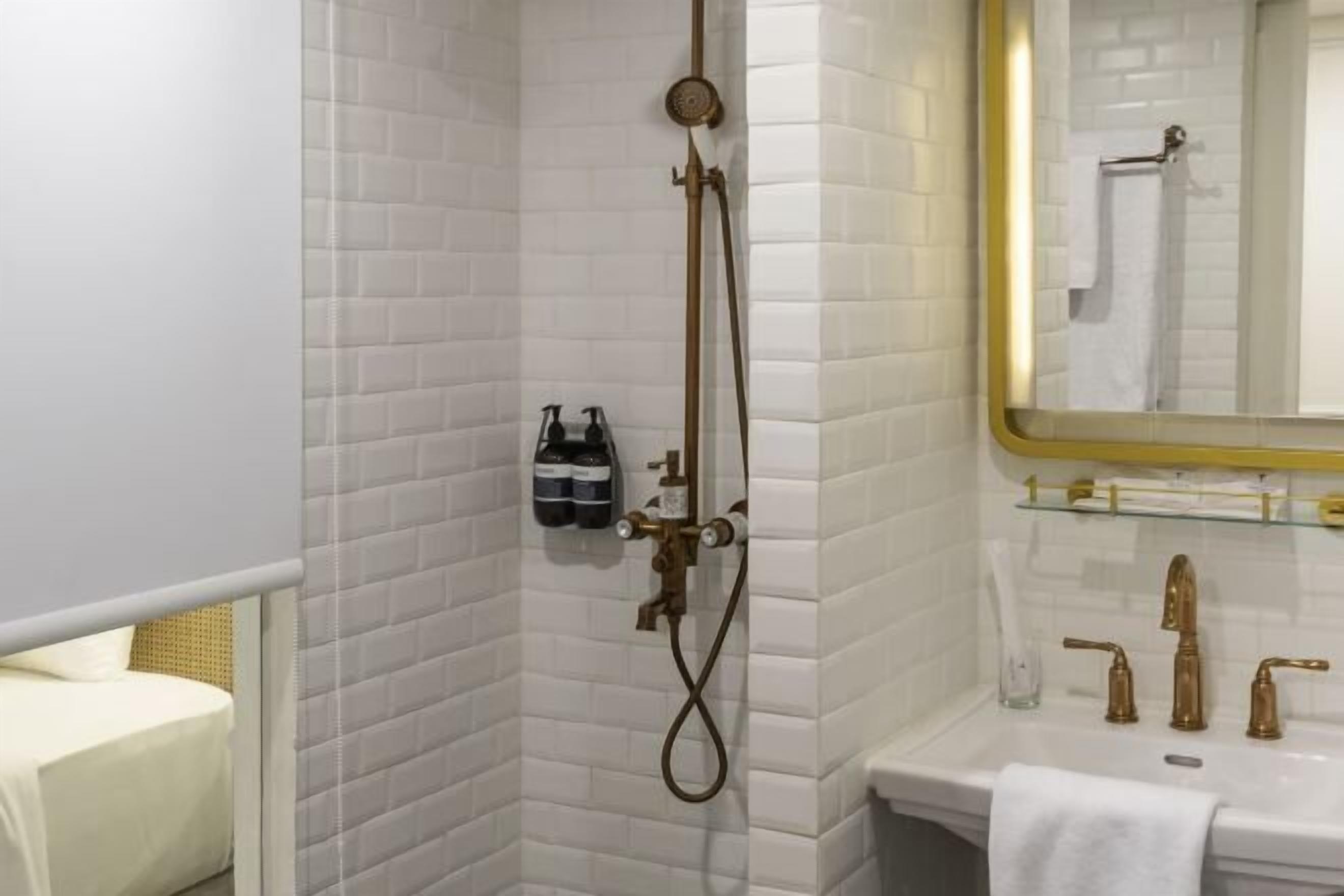 A bathroom with white tiled walls, a shower with a gold showerhead, a sink with gold faucets, and a mirror with a gold frame.