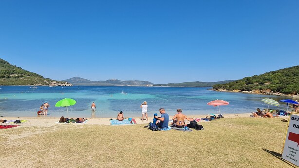 A beach with people sunbathing and swimming, a sign for a beach bar, and a clear view of the sea and hills in the background. - Elegant deluxe cottage close to beaches (Sardegna)