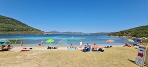 A beach with people sunbathing and swimming, a sign for a beach bar, and a clear view of the sea and hills in the background.