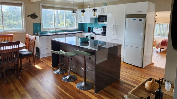 A modern kitchen with a central island, bar stools, a dining area with a wooden table and chairs, and a view of the outdoors through large windows. - # 40 At the Beach (Waitārere Beach)