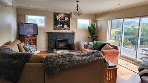 A living room with a fireplace, sofa, television, and large windows offering a view of the outdoors. - # 40 At the Beach (Waitārere Beach)