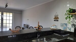 Cocina moderna con encimera de granito negro, pileta de acero inoxidable y una olla negra sobre la encimera. Se ve una ventana con persianas, una planta a la derecha y una máquina de café de pared.