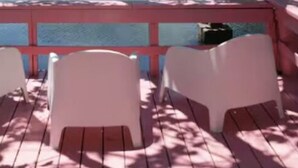 A wooden deck with a red railing and three white chairs.