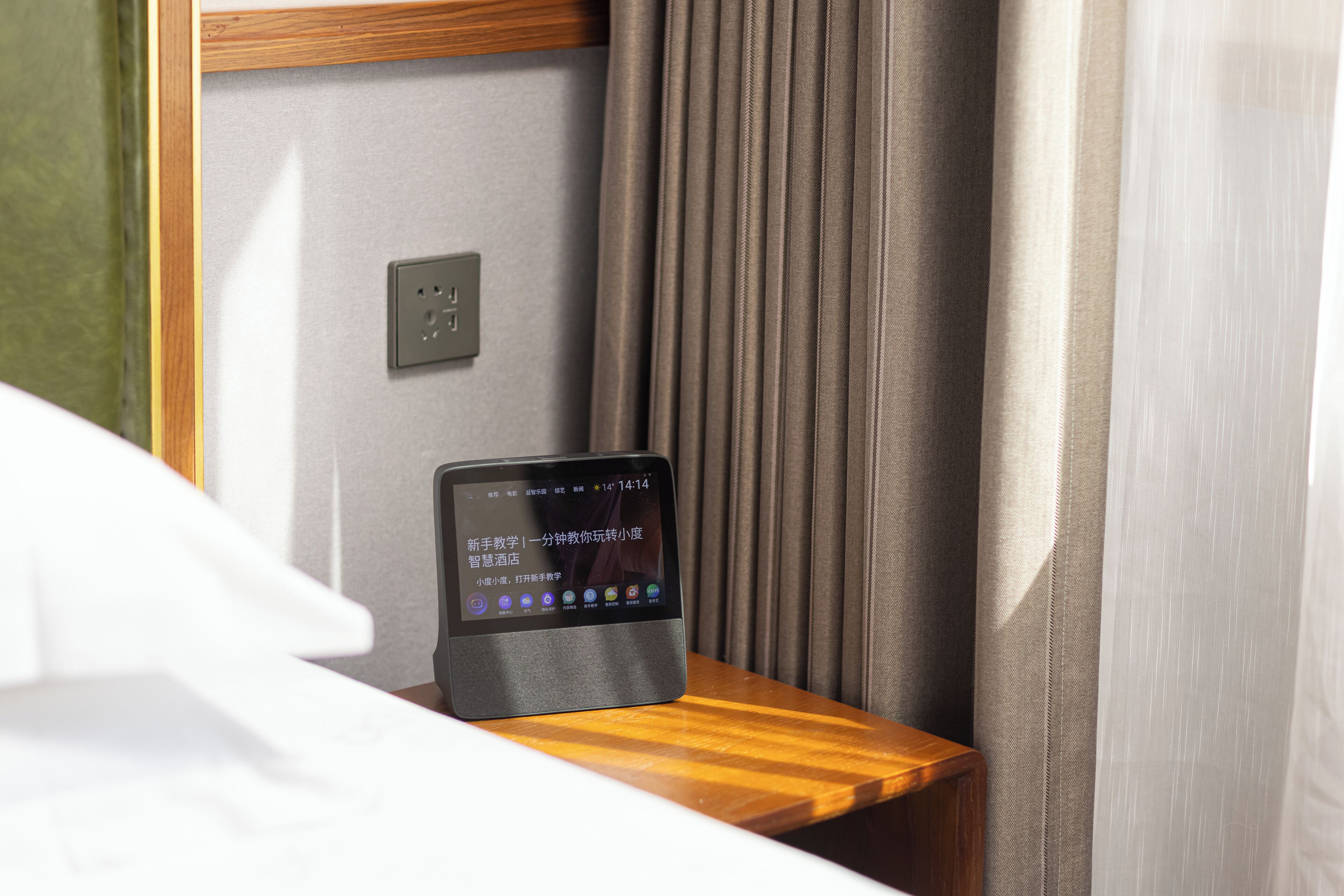 A tablet on a wooden nightstand in a hotel room, with a curtain and a wall socket visible.
