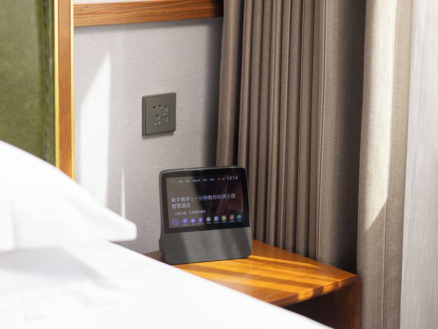 A tablet on a wooden nightstand in a hotel room, with a curtain and a wall socket visible.