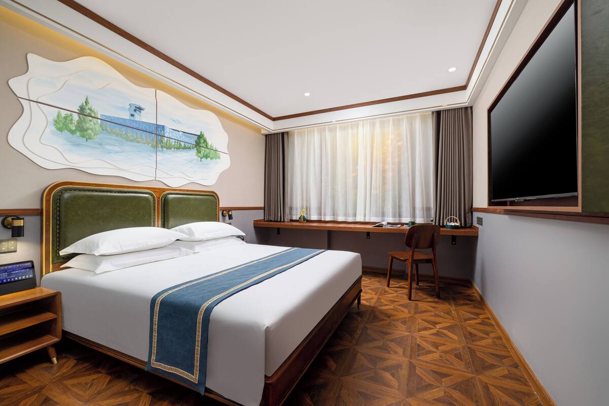 A hotel room with a large bed, a desk, a chair, a TV, and a painting on the wall.