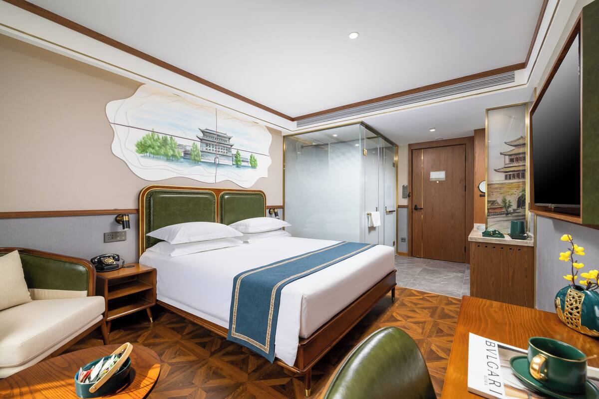 A hotel room with a bed, a desk, a chair, a sofa, a TV, and a painting on the wall.